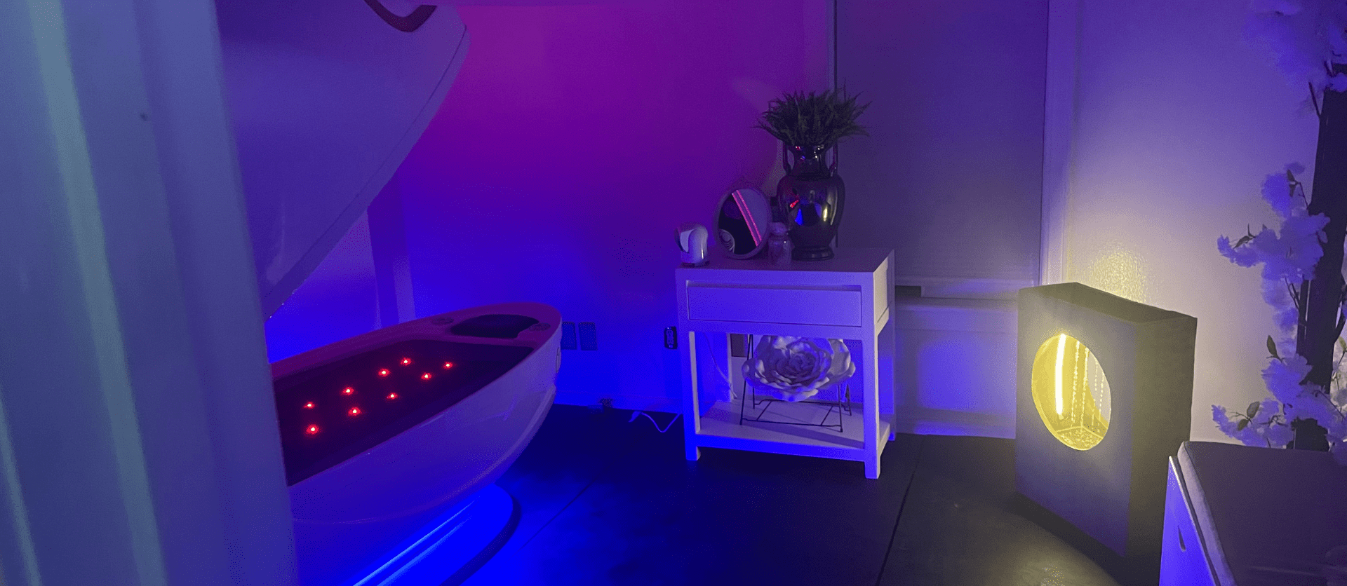 A serene spa room glows with calming blue and purple lighting, featuring a Cocoon Wellness Pod illuminated with red lights.