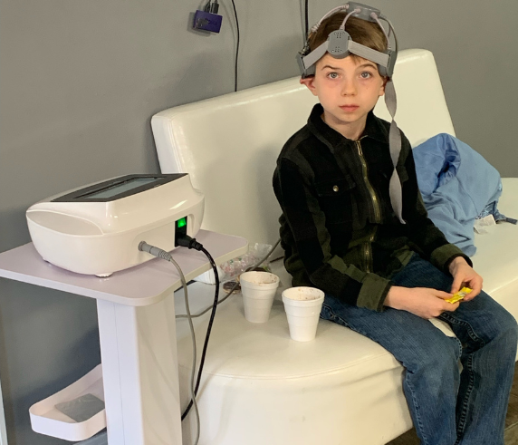 Young client receiving a Repetitive Transcranial Magnetic Stimulation (rTMS) session at Heat and Wellness Spa in Ripley, MS using a non-invasive magnetic stimulation device to support brain wellness, focus, and cognitive function.